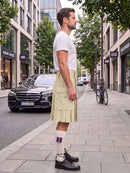 A man stands on a city sidewalk wearing a white t-shirt, a light beige pleated kilt, white socks with purple stripes, and black leather shoes. A black Mercedes-Benz car is parked on the street behind him, with trees and storefronts in the background.