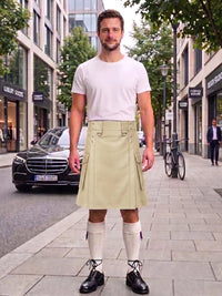 A man stands on a city sidewalk wearing a light beige pleated kilt with a white t-shirt, white knee-high socks, and black shoes. A black Mercedes-Benz car and a bicycle are visible in the background of the street scene.