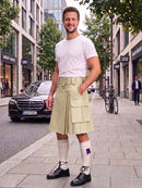 A man wearing a light beige kilt with front flap pockets, a white t-shirt, white knee-high socks, and black leather shoes, standing on a city sidewalk.