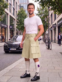A man wearing a light beige kilt with front flap pockets, a white t-shirt, white knee-high socks, and black leather shoes, standing on a city sidewalk.