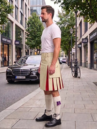 A man wearing a beige kilt with red pleated accents, a white t-shirt, white knee-high socks, and black leather shoes, standing on a city sidewalk next to a street with cars and trees.