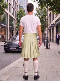 A man standing on a city sidewalk, viewed from behind, wearing a white t-shirt and a light olive green pleated kilt with white knee-high socks and black shoes.