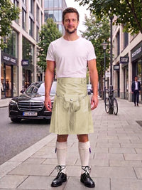 A man stands on a city sidewalk wearing a white t-shirt, a light olive green kilt with a front pouch pocket, white knee-high socks, and black leather shoes. A black Mercedes-Benz car and city buildings are visible in the background.
