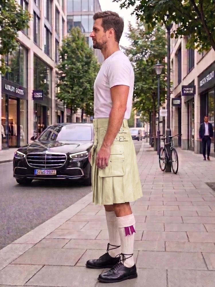 A man standing on a city sidewalk wearing a pale yellow-green pleated kilt with embroidered details, a white t-shirt, white socks with a purple bow, and black shoes. A black Mercedes-Benz car and urban buildings are visible in the background.