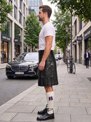 A man standing on a city sidewalk wearing a black leather kilt, white t-shirt, white socks, and black shoes, with a street scene and parked cars in the background.