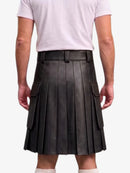 A person shown from the back wearing a black pleated leather kilt with side cargo pockets, paired with a light pink t-shirt and white socks, against a plain light gray background.