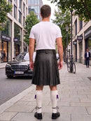 A man viewed from behind wearing a black leather kilt, white t-shirt, white knee-high socks with purple tassels, and black shoes, standing on a city sidewalk.