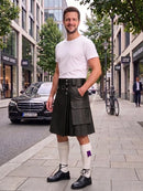 A man stands on a city sidewalk wearing a black leather kilt with a matching belt, a white t-shirt, white knee-high socks, and black leather shoes. A black Mercedes-Benz car and urban buildings are visible in the background.