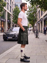A man standing on a city sidewalk wearing a dark green leather kilt, white t-shirt, white knee-high socks with a purple ribbon, and black leather shoes. A black Mercedes-Benz car and a bicycle are parked on the street behind him.
