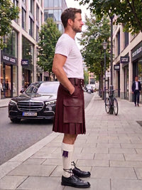 A man standing on a city sidewalk wearing a dark green pleated kilt, a white t-shirt, white socks, and black leather shoes. A black Mercedes-Benz car is parked on the street behind him, with trees and storefronts in the background.