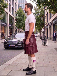 A man standing on a city sidewalk wearing a dark burgundy leather kilt, white t-shirt, white socks, and black shoes, with a street scene and parked cars in the background.