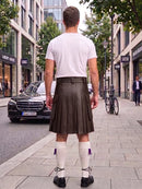 A man standing on a city sidewalk, viewed from behind, wearing a white t-shirt and a dark brown pleated leather kilt with white knee-high socks and black shoes.