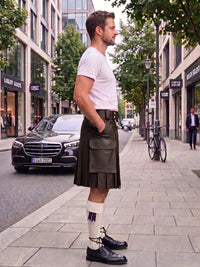 A man standing on a city sidewalk wearing a dark green leather kilt, white t-shirt, white knee-high socks with a purple ribbon, and black leather shoes. A black Mercedes-Benz car and a bicycle are parked on the street behind him.