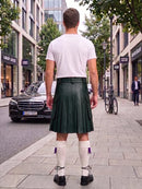 A man standing on a city sidewalk, viewed from behind, wearing a dark green pleated leather kilt, a white t-shirt, white knee-high socks, and black shoes.