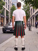 A man viewed from behind wearing a dark green pleated leather kilt with red vertical stripes, a white t-shirt, white knee-high socks, and black shoes, standing on a city sidewalk.