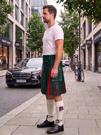 A man wearing a black leather kilt with red pleats, a white t-shirt, white knee-high socks, and black leather shoes stands on a city sidewalk next to a street with cars and trees.