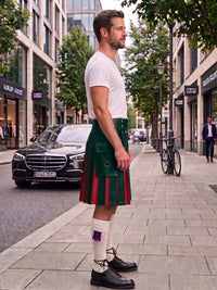 A man standing on a city sidewalk wearing a green kilt with red and white stripes, a white t-shirt, white socks, and black leather shoes. The background shows a street with buildings, a parked black car, and a bicycle.