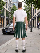A man standing on a city sidewalk, photographed from behind, wearing a dark green leather kilt with white vertical stripes, a white t-shirt, white knee-high socks, and black shoes. Storefronts, trees, and a parked black car are visible in the background.