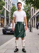 A man stands on a city sidewalk wearing a dark green kilt with white geometric stripe details, a white t-shirt, white knee-high socks, and black leather shoes. The background shows a blurred urban street scene with buildings, a parked black car, and a bicycle.