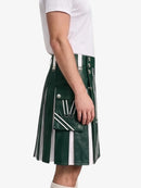 A side view of a person wearing a dark green pleated faux-leather kilt with white vertical stripes, white piping, and silver-tone metal hardware, paired with a white t-shirt.