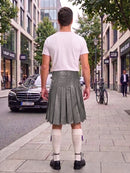 A man standing on a city sidewalk, viewed from behind, wearing a white t-shirt and a dark gray pleated leather kilt with white knee-high socks and black shoes. Storefronts, trees, and a parked black car are visible in the background.