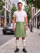 A man stands on a city sidewalk wearing a white crew-neck t-shirt and an olive green pleated leather kilt with silver hardware, paired with white knee-high socks and black leather shoes.