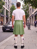 A man standing on a city street, viewed from behind, wearing a white t-shirt, a light green pleated kilt, white knee-high socks, and black shoes.