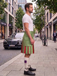 A man standing on a city sidewalk wearing a green kilt with red and purple pleats, a white t-shirt, white knee-high socks, and black leather shoes. A black Mercedes-Benz car is parked on the street behind him.