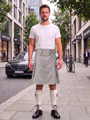 A man stands on a city sidewalk wearing a light gray pleated kilt, a white t-shirt, white knee-high socks, and black leather shoes. The background shows a street with buildings, a parked black Mercedes, and a bicycle.