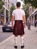 A man standing on a city sidewalk, viewed from behind, wearing a white t-shirt and a dark brown pleated faux-leather kilt with white knee-high socks and black shoes.