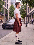 A man standing on a city sidewalk wearing a dark brown leather kilt, a white t-shirt, white socks, and black leather shoes, with a street scene and parked cars in the background.
