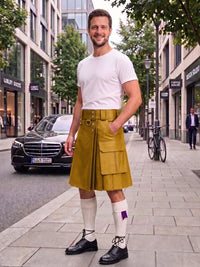 A man stands on a city sidewalk wearing a mustard yellow kilt with a matching belt and buckle, a white t-shirt, white knee-high socks, and black shoes. A black Mercedes-Benz car is parked on the street behind him, with trees and storefronts in the background.