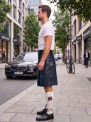 A man standing on a city sidewalk wearing a white crew-neck t-shirt, a dark navy blue leather kilt, white socks with a purple bow, and black leather shoes. A black Mercedes-Benz car is parked on the street behind him.
