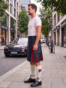 A man wearing a black leather kilt with red pleats, a white t-shirt, white knee-high socks, and black leather shoes stands on a city sidewalk next to a street with cars and trees.