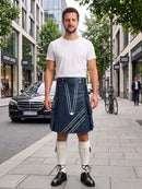 A man stands on a city sidewalk wearing a white crew-neck t-shirt and a dark navy blue kilt with white geometric line patterns. He is also wearing white knee-high socks and black leather shoes.