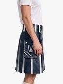 A model wearing a navy blue leather pleated kilt with white vertical stripes and silver-tone hardware, paired with a white t-shirt against a light gray background.