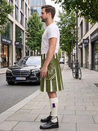 A man standing on a city sidewalk wearing a white t-shirt, a green pleated kilt with white stripes and a white 'W' logo, white knee-high socks, and black leather shoes. A black Mercedes-Benz car and a bicycle are parked on the street behind him.