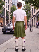 A man standing on a city sidewalk, viewed from behind, wearing an olive green pleated kilt with a white t-shirt, white knee-high socks, and black shoes. Buildings, trees, and parked cars are visible in the background.