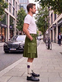 A man standing on a city sidewalk wearing a green pleated kilt, white t-shirt, white knee-high socks, and black leather shoes, with buildings, trees, and parked cars in the background.
