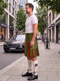 A man standing on a city sidewalk wearing a green pleated leather kilt with red and purple striped accents, a white t-shirt, white knee-high socks, and black leather shoes. A black Mercedes-Benz is parked on the street behind him.