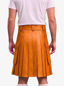 A person shown from the back wearing a mustard-orange pleated leather kilt with side cargo pockets, paired with a light pink t-shirt and white socks, against a plain light gray background.