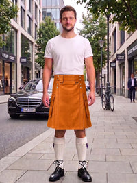 A man standing on a city sidewalk wearing a mustard yellow corduroy kilt, a white t-shirt, white knee-high socks, and black leather shoes. The background shows a street with buildings, a parked black Mercedes, and a bicycle.