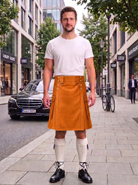 A man stands on a city sidewalk wearing a mustard-orange kilt, white t-shirt, white knee-high socks, and black leather shoes. The background shows a street with buildings, a parked black Mercedes, and a bicycle.