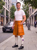 A man standing on a city sidewalk wearing a bright orange-brown leather kilt with a matching belt, a white t-shirt, white knee-high socks, and black leather shoes. A Mercedes-Benz car and urban buildings are visible in the background.