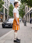 A man stands on a city sidewalk wearing a bright orange kilt with white vertical stripe details, a white t-shirt, cream knee-high socks, and black leather dress shoes. Modern buildings, trees, a parked black Mercedes, and a bicycle are visible in the background.