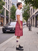 A man standing on a city sidewalk wearing a white t-shirt, a dusty rose pleated kilt, white socks with purple stripes, and black leather shoes. A black Mercedes-Benz car is parked on the street behind him.