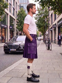 A man standing on a city sidewalk wearing a dark green pleated kilt, a white t-shirt, white socks, and black leather shoes. A black Mercedes-Benz car is parked on the street behind him, with trees and storefronts in the background.