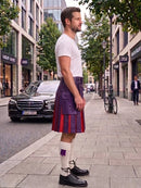 A man standing on a city sidewalk wearing a purple and red pleated kilt, a white t-shirt, white socks, and black leather shoes, with buildings, trees, and parked cars in the background.