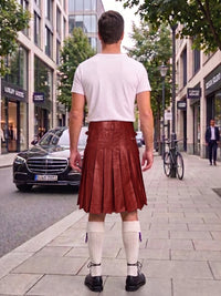 A man standing on a city sidewalk, viewed from behind, wearing a white t-shirt and a reddish-brown pleated leather kilt with white knee-high socks and black shoes.