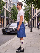 A man standing on a city sidewalk wearing a white t-shirt, a blue leather kilt, white socks with purple accents, and black leather shoes. A black Mercedes-Benz car is parked on the street behind him, with trees and storefronts in the background.